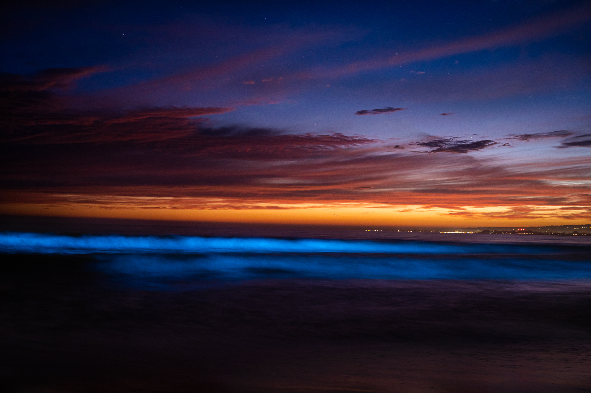 Purple, orange, and blue skies dim over a glowing blue wave.