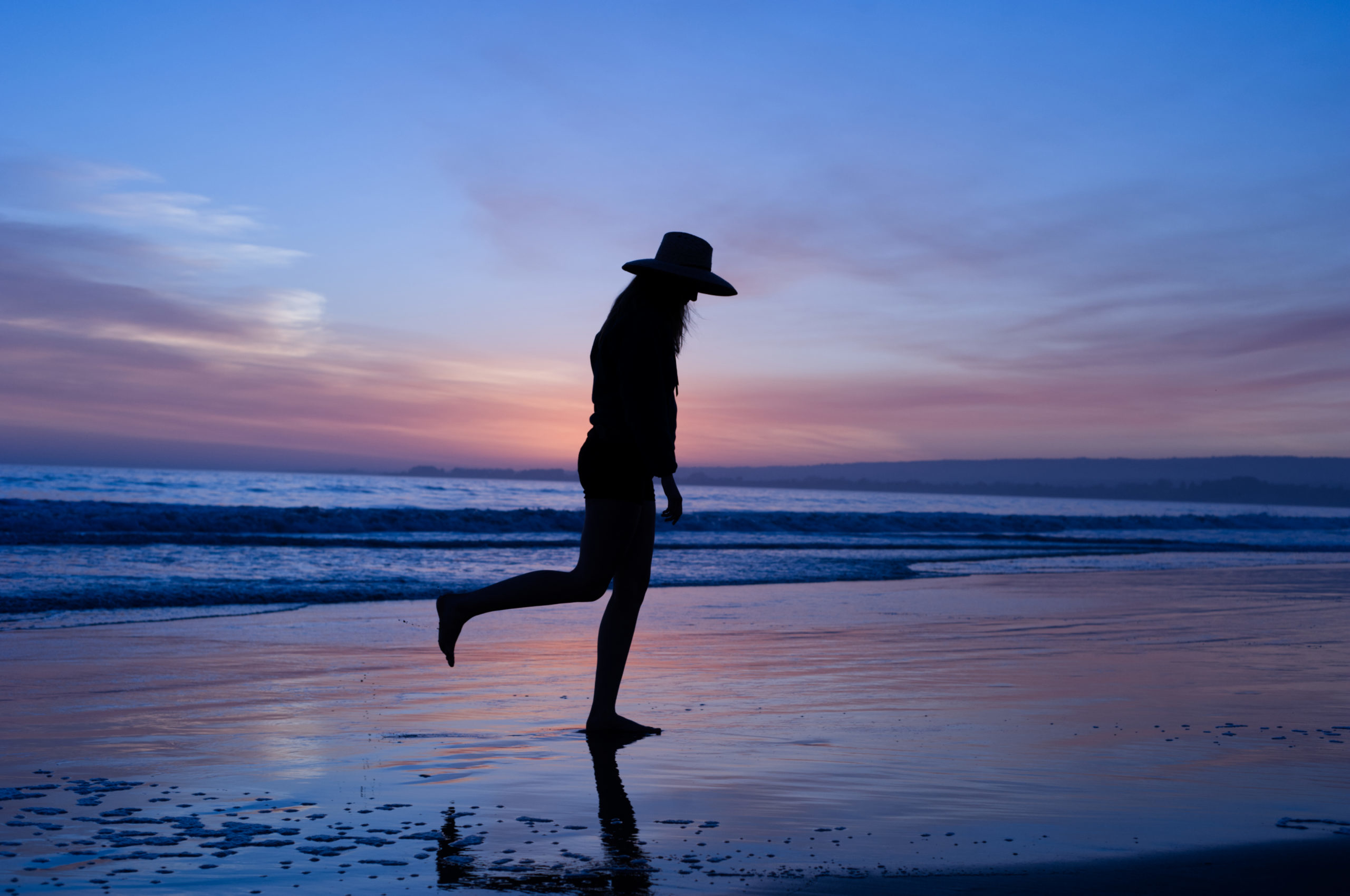 The silhouette of a woman in a hat walking along the beach at dusk.