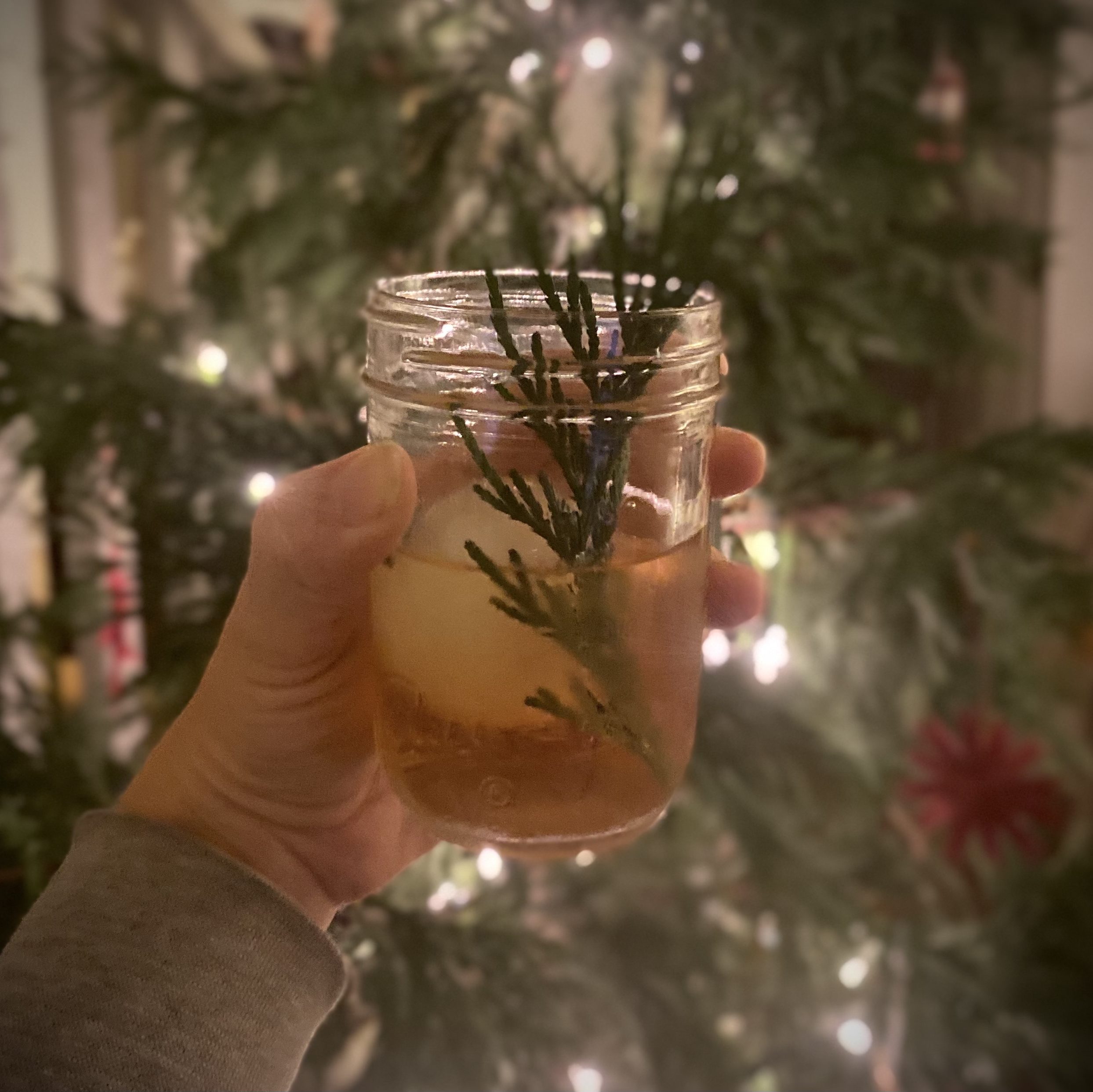 A cocktail in a jar garnished with a sprig of incence cedar leaves in front of a decorated tree.