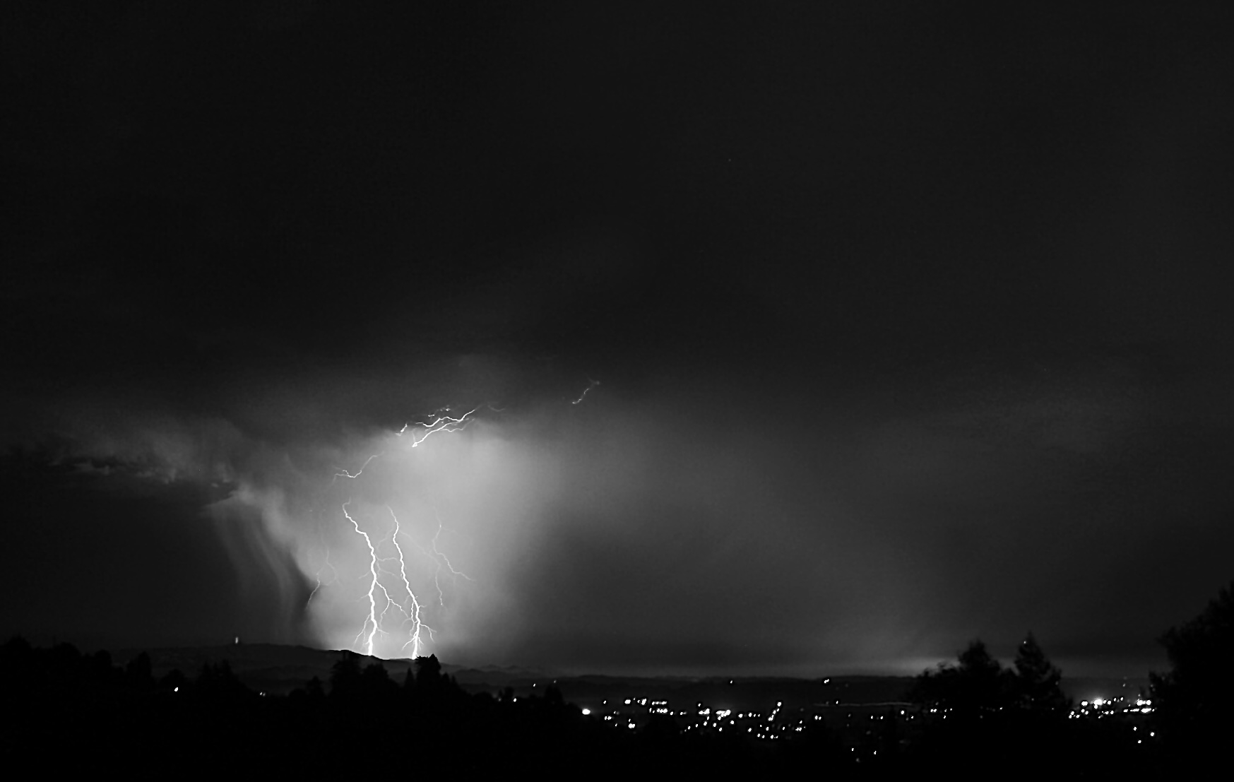 A black and white image of lightening (left) over some city lights.