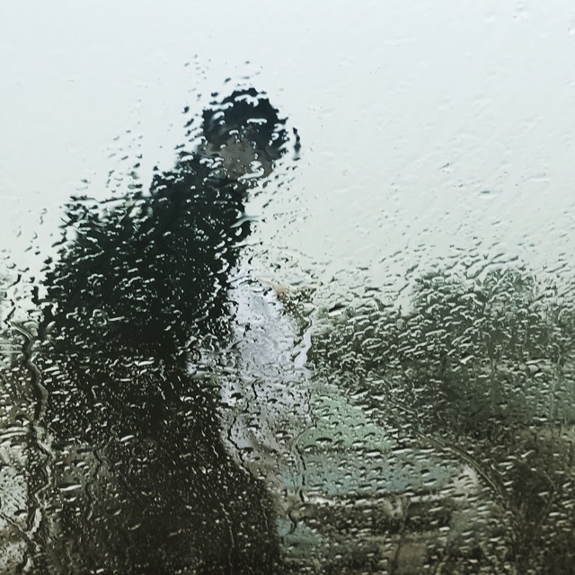 The silhouette of a figure walking past cars is obscured with rain on a glass window.