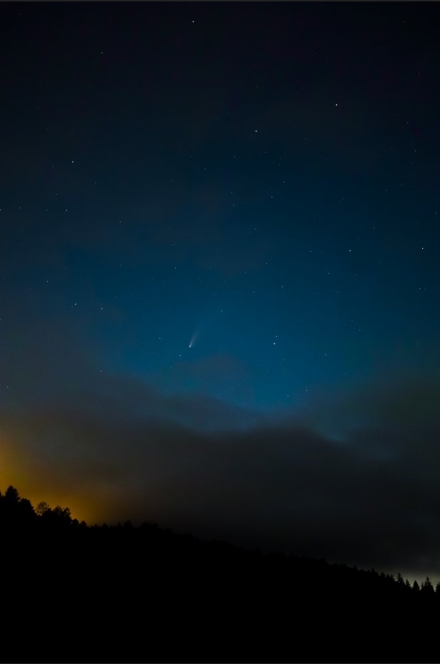 A small white streak smudges across a blue night sky with the orange glow of the city peaking over a mountain silhouette.