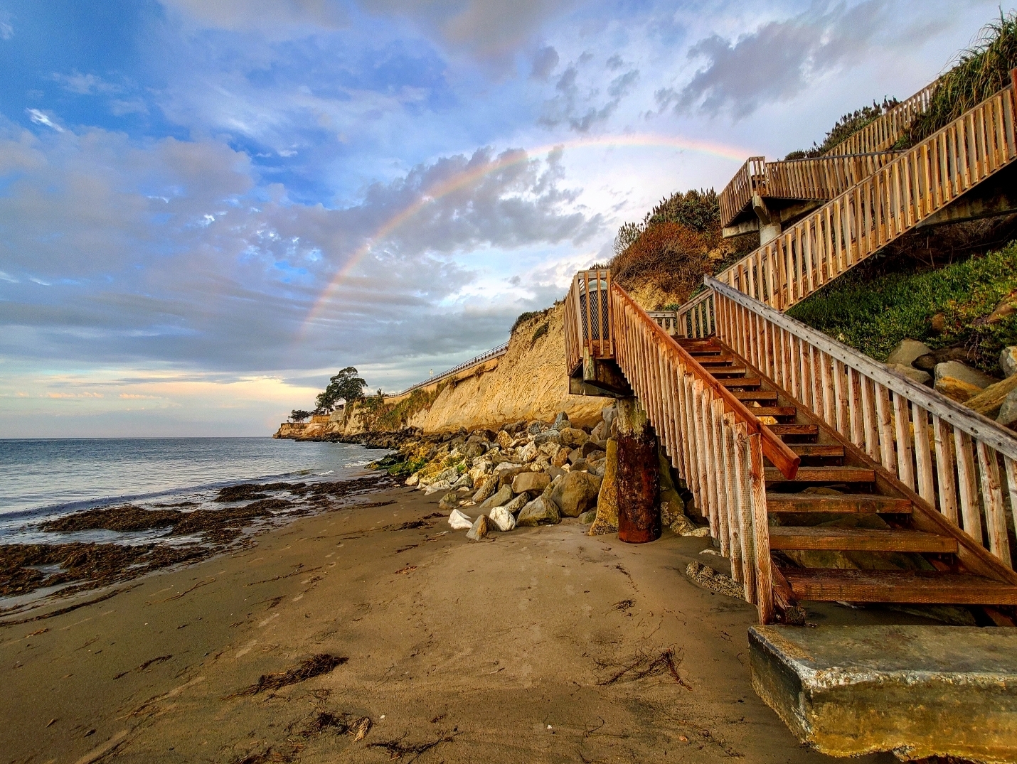 Stairs lead off of a beach with a rainbow in the sky behind.