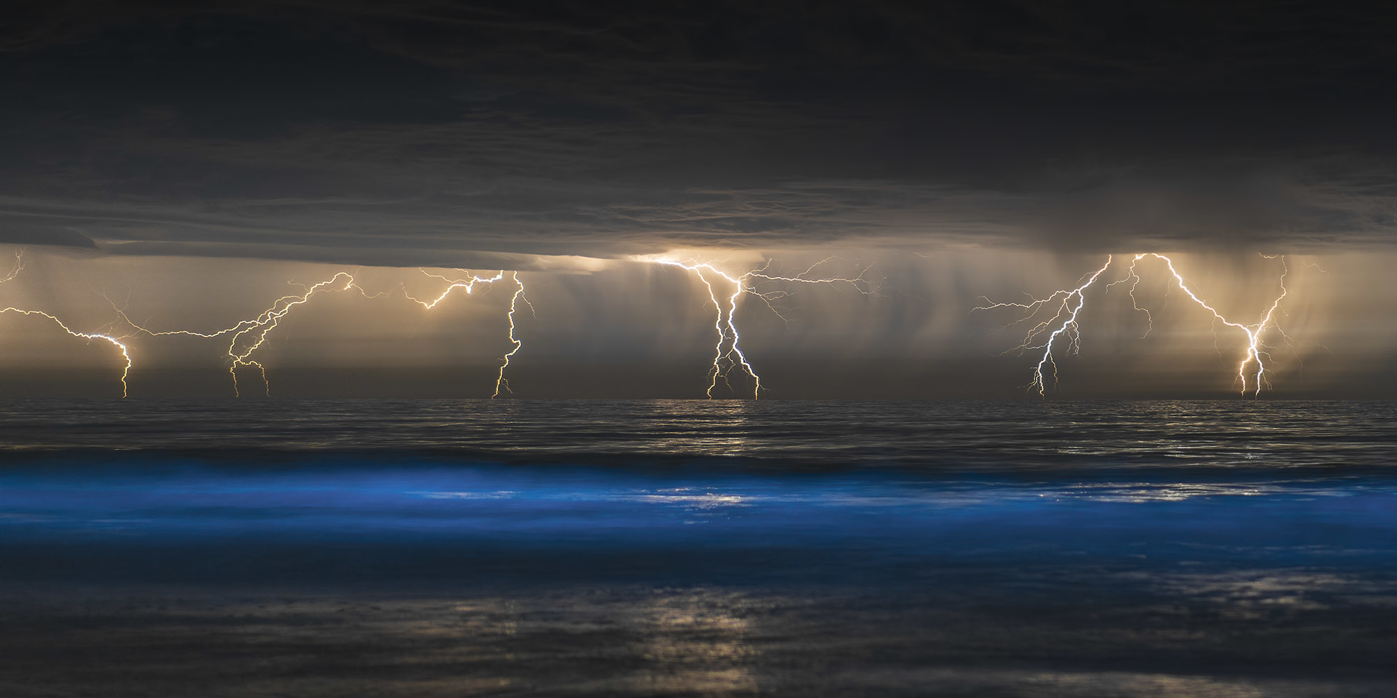 A series of lightening bolts descends grey clouds towards the ocean, with a cresting wave of glowing blue in the foreground.