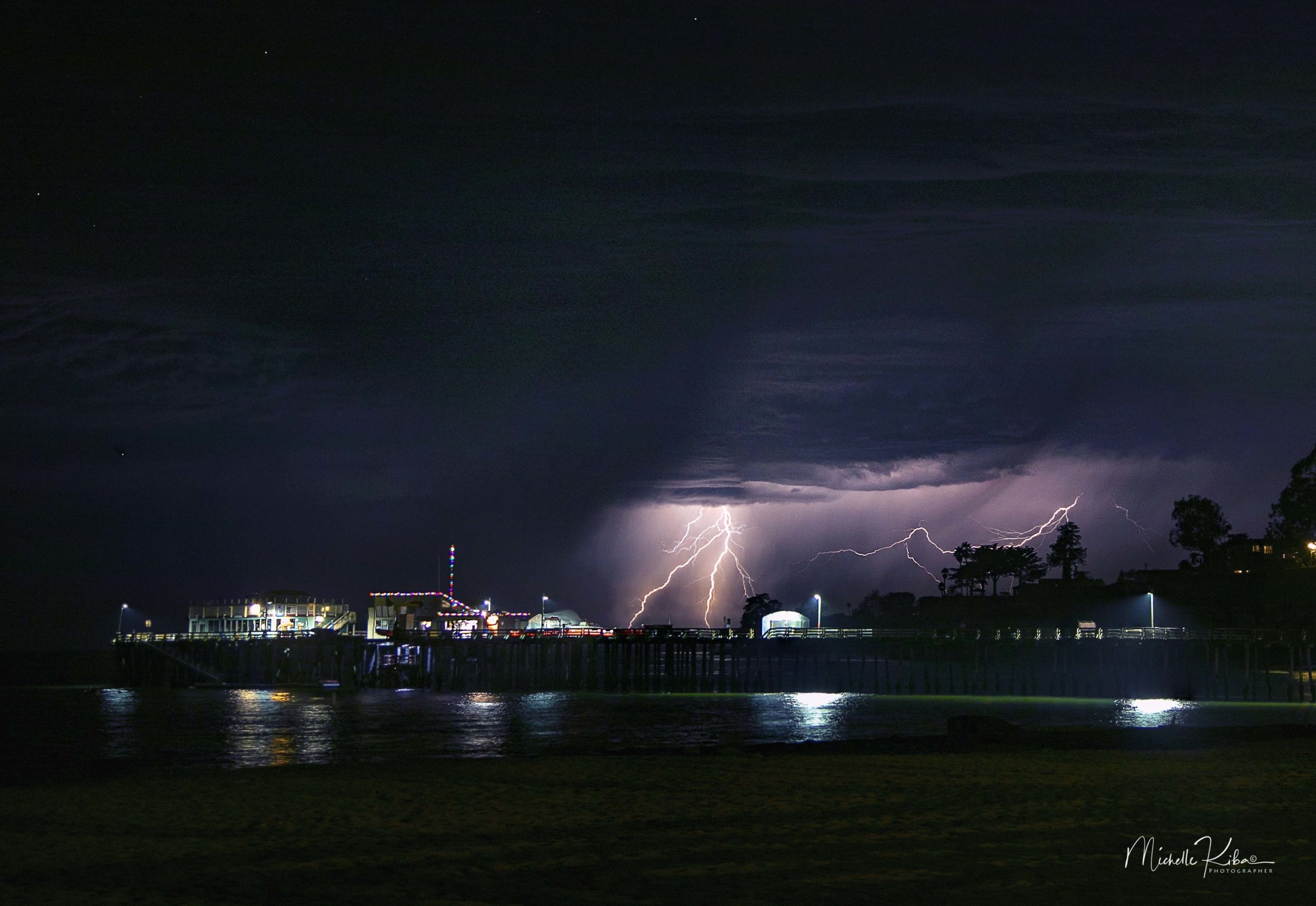 Lightning flashes in a purple sky behind the Capitola wharf at night.