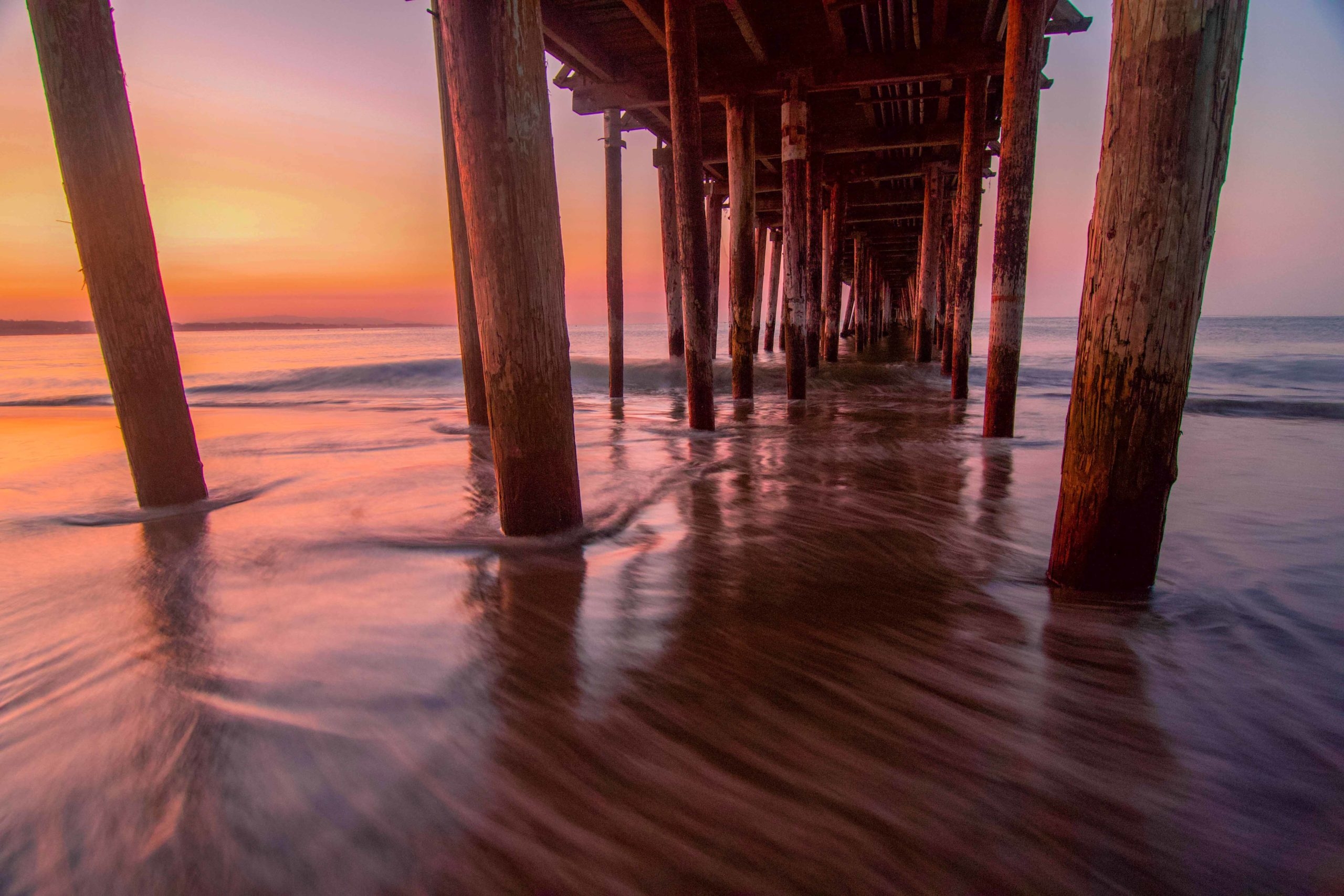 The pillars of the pier extend out into the ocean on both sides, with soft waves under a purple and orange sky.