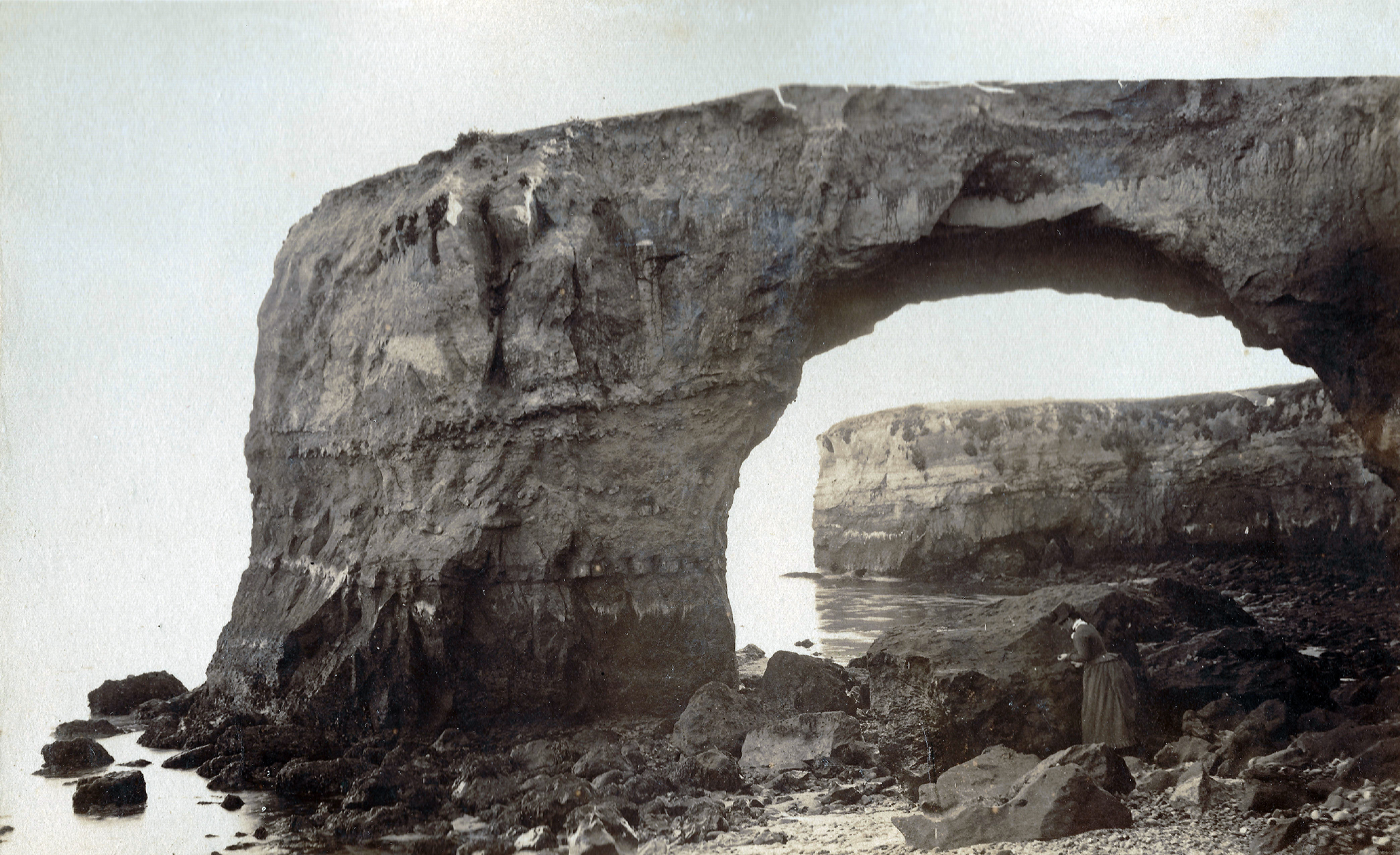 A black and white photograph of a woman exploring the tidepool below a natural bridge rock formation.