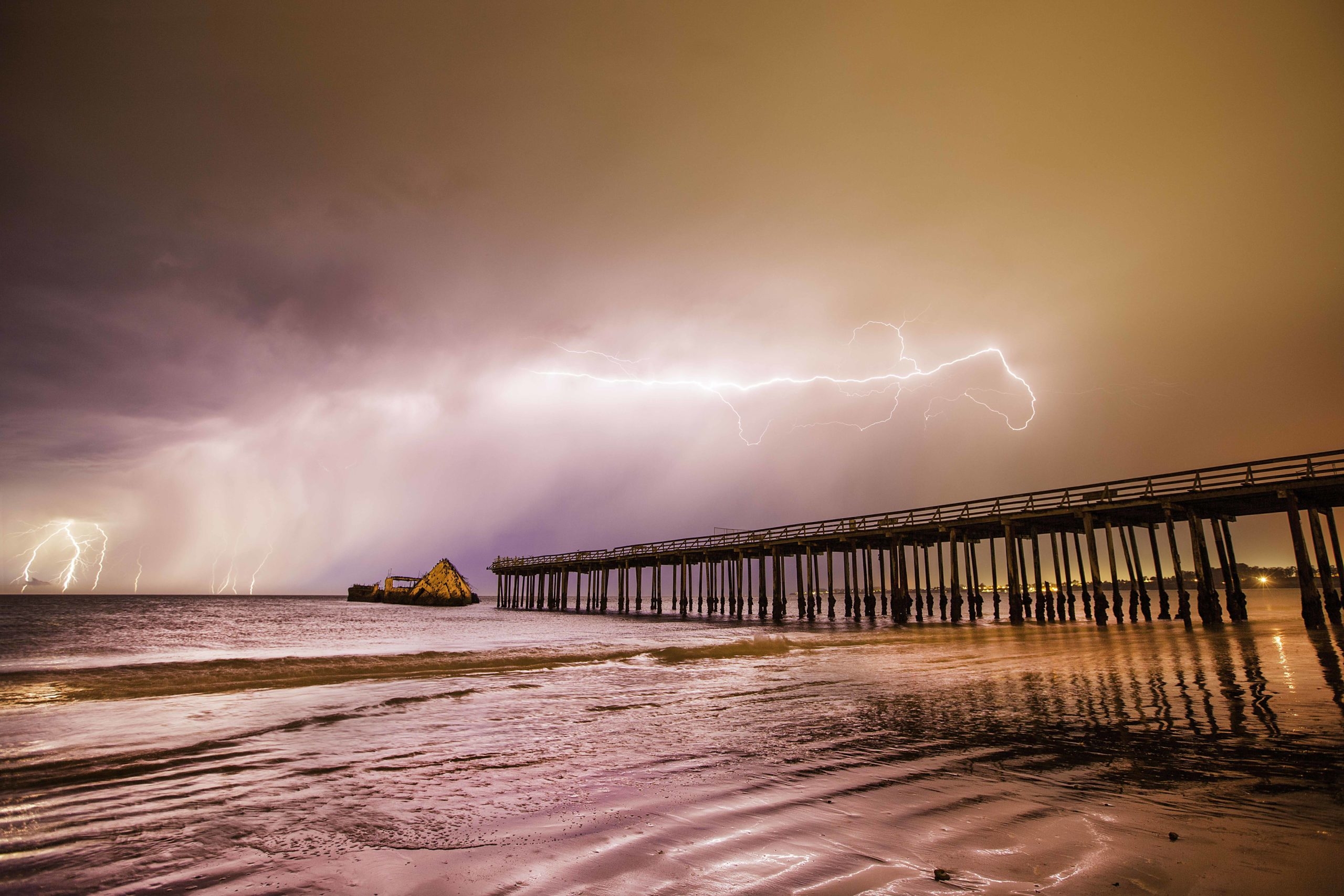 A bright purple and pink view of the cement ship at the end of a pier with a bolt of lightening creeping across the sky above.