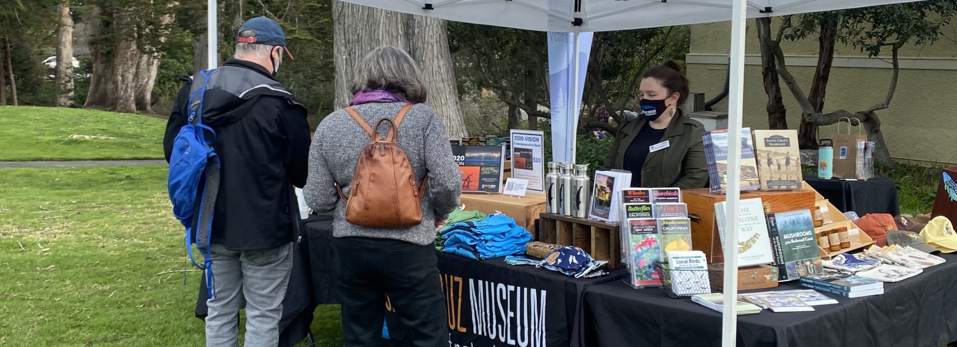 Two people explore tables full of merchandise under a pop-up tent outside, while a masked staff person looks on.